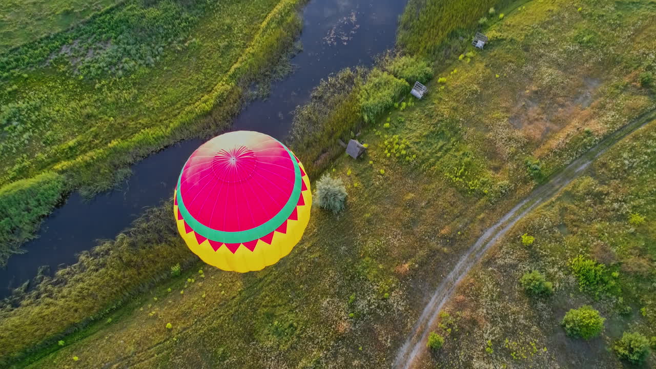 Aerostat on nature background. Colorful hot air balloon flying over the field near the small river. Outdoor activity. Top view. Camera moves down.