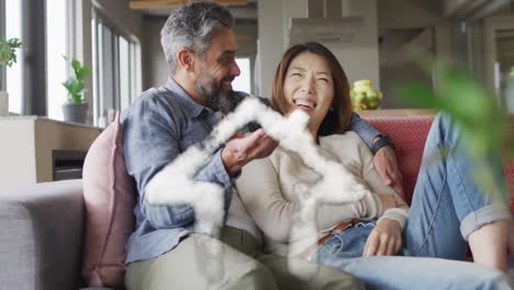 Animation of house shape over diverse couple embracing on sofa at home