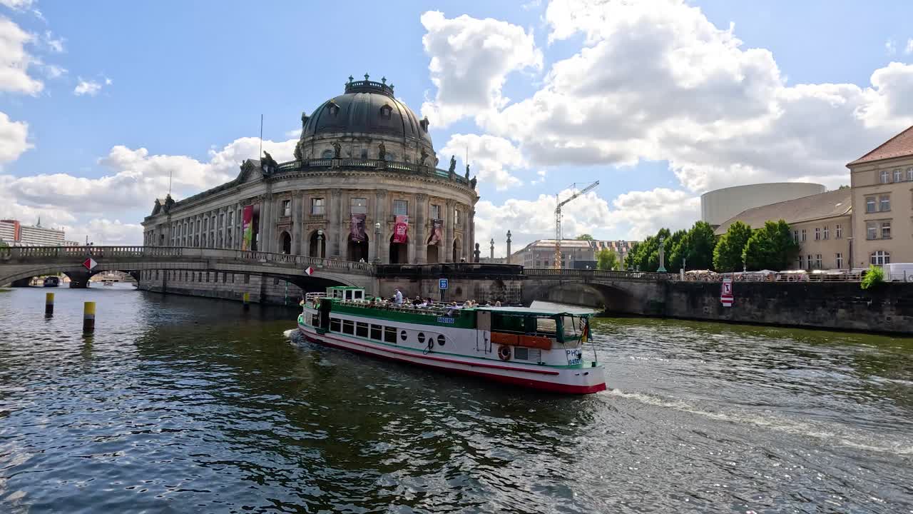A passenger tour boat glides along the Spree River past a grand historic museum under bright daylight, with dynamic water reflections and a wide-angle view