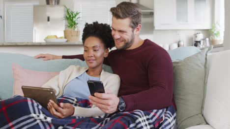 video of happy diverse couple embracing and using electronic devices on sofa