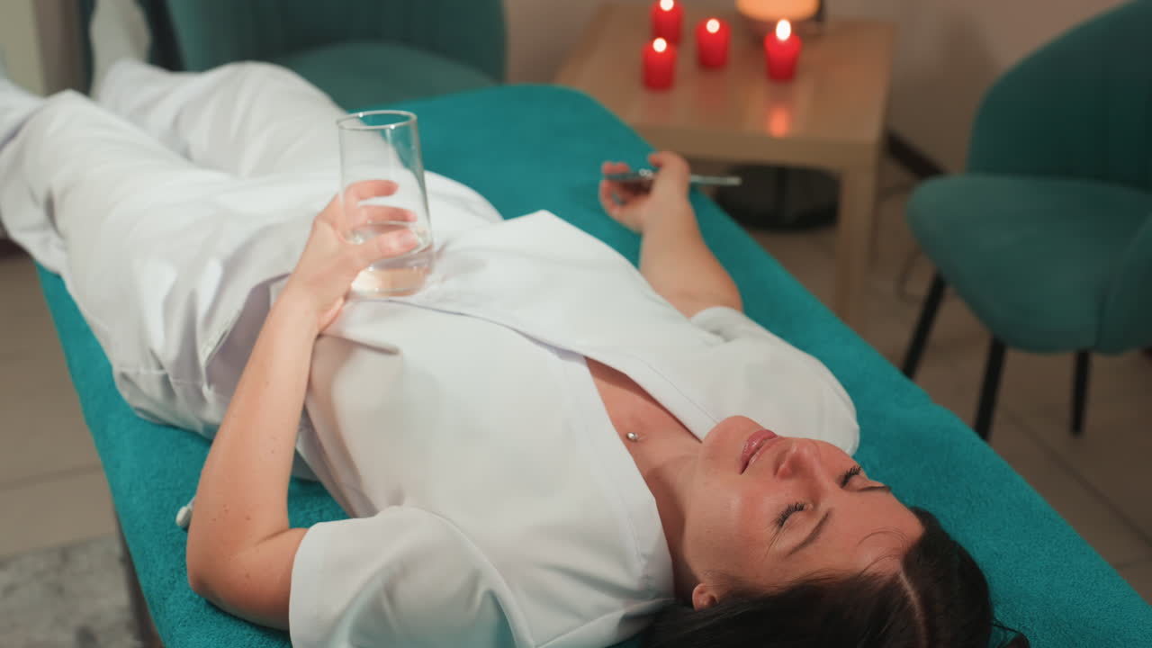 Midaldrende hvid kvinde ligger på sengen og får blid mikrostømsmassage, afslappet udtryk, holder et glas vand, hvid badekåbe, turkis behandlingsbord, levende lys-atmosfære, klinikstol i baggrunden.