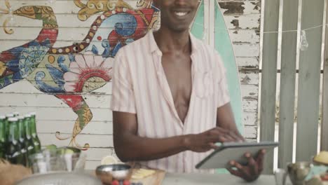 portrait of happy african american barman using tablet behind the counter at beach bar, slow motion