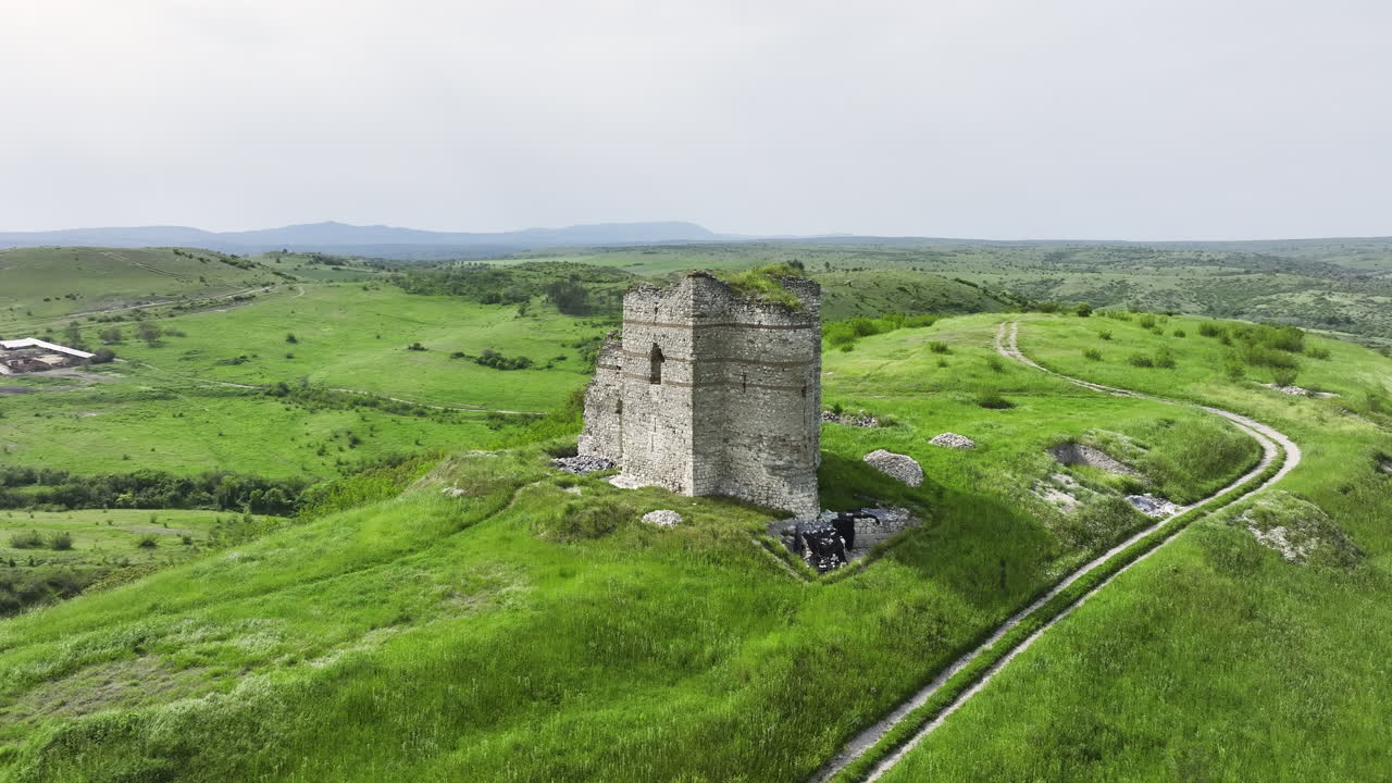 POI drone orbit of a hilltop medieval fortress with a visible footpath, lush spring greenery and overcast skies over rolling countryside in Bulgaria