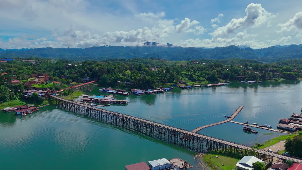 Wide aerial orbit of Mon Bridge in Sangkhlaburi, Thailand. The view shows the wooden structure, floating houses, lake, green hills, and distant mountains under partly cloudy skies