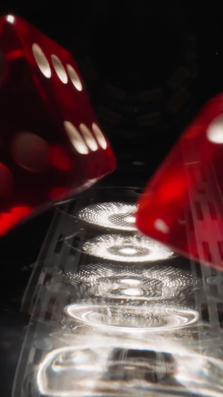 Red dices with white dots thrown into empty glass hitting on sides at bright light. Small cubes for gambling games rotate in wineglass on black background macro