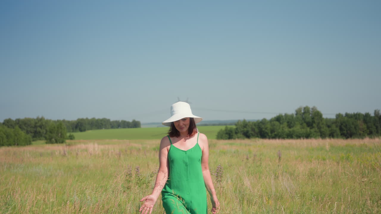 lady walking through tall grass wearing green summer gown and white hat as she gently touches grass blades in meadow with joyful expression under clear blue sky and tree line