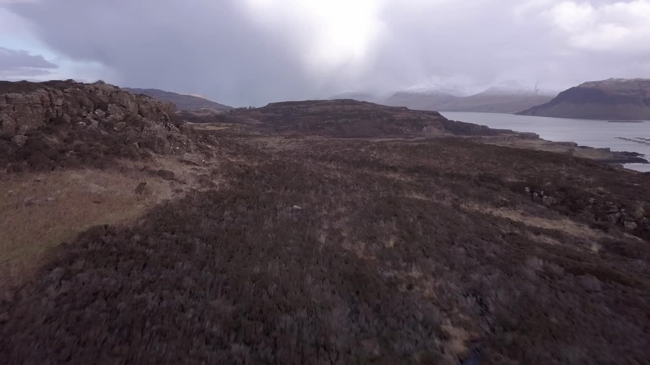 Aerial shot flying low and slow in an easterly direction towards snowcapped hills on Mull, from the island of Ulva, over peaches of heather