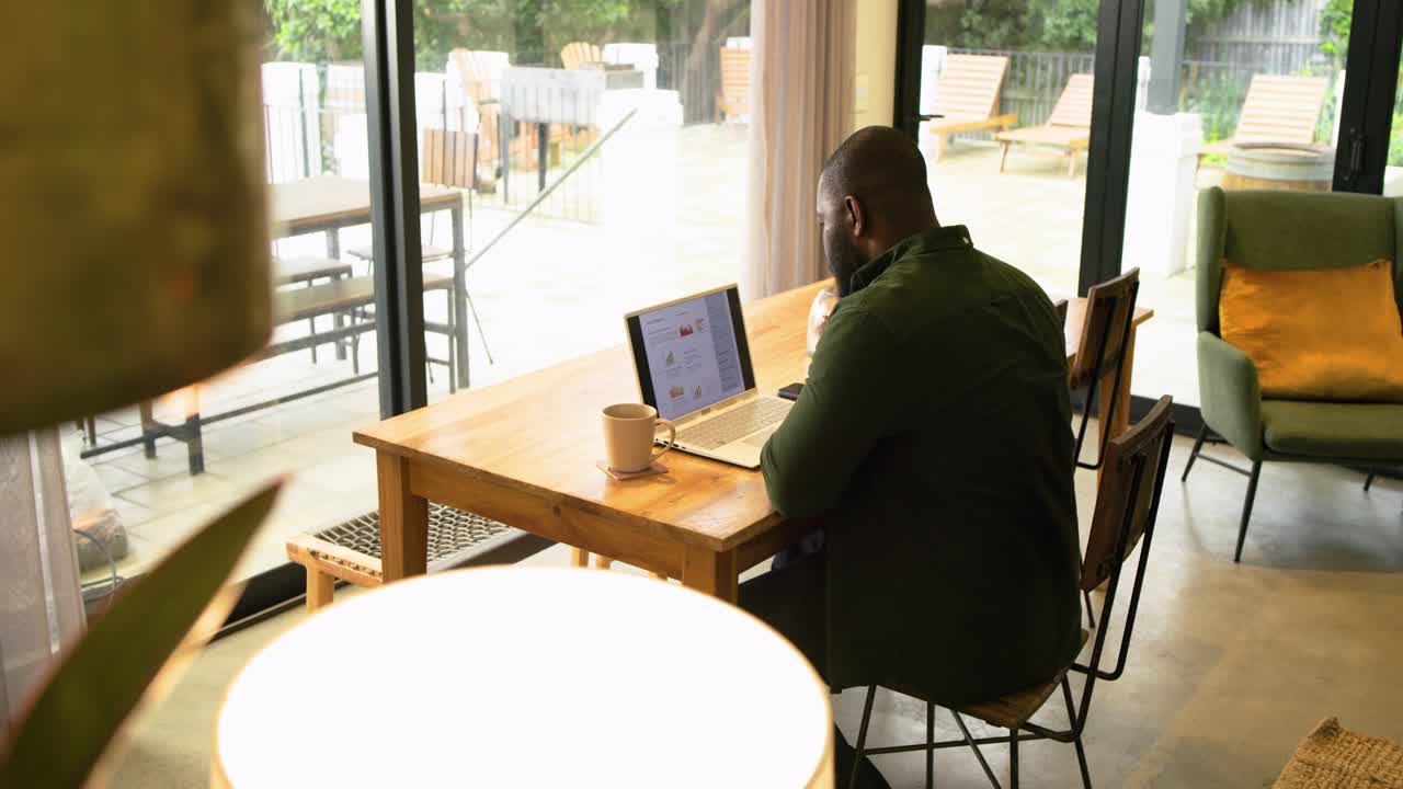 Man is opening laptop to work, scrolling screen, snacking fruit, sipping coffee near glass doors