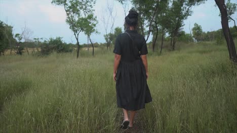 a girl with combed hair in a long black dress is walking along a path in tall grass, which she is stroking with her hand