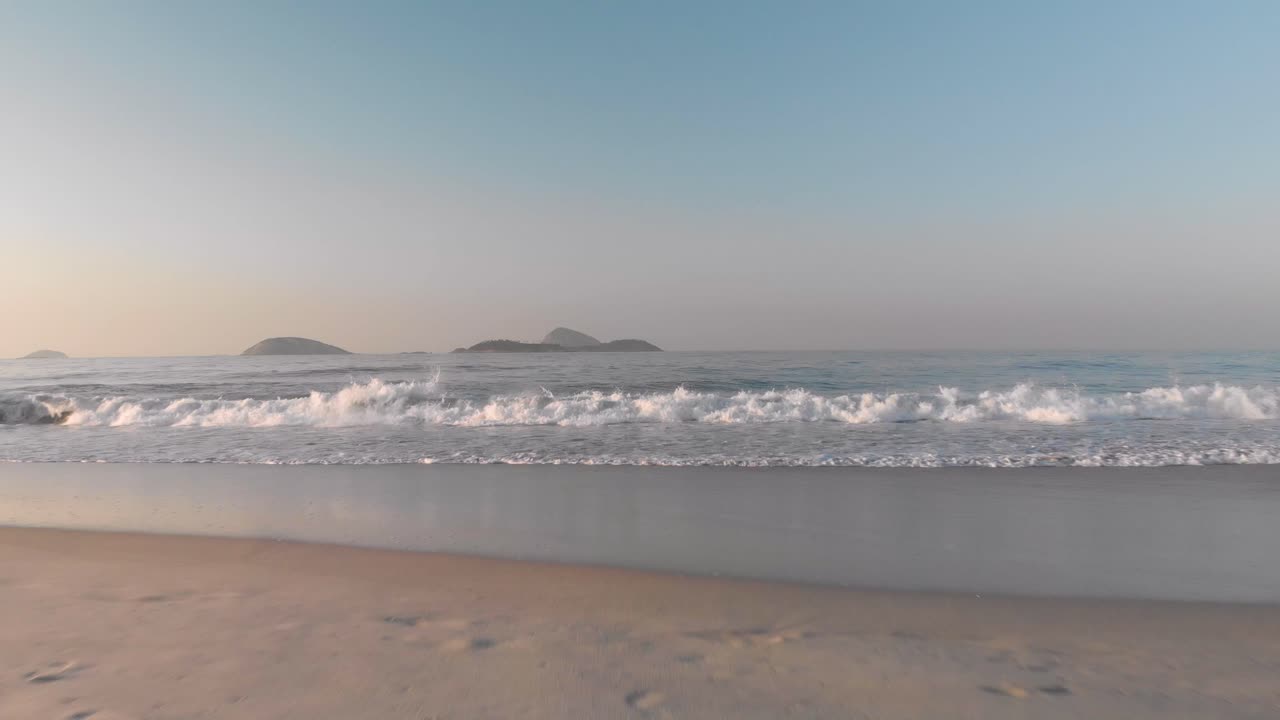 temprano en la mañana, vista aérea baja del océano tranquilo con pequeñas olas rompiendo desde la playa con huellas acercándose y flotando sobre el agua al amanecer con islas al fondo.