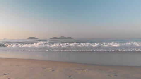 Early-morning-low-aerial-view-of-the-calm-ocean-with-small-breaking-waves-starting-from-the-beach-with-footprints-approaching-and-hovering-over-the-water-at-sunrise-with-islands-in-the-background