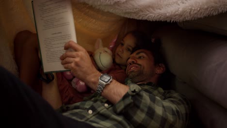 un hombre moreno en una camisa a cuadros verde junto con su hija pequeña, una niña morena en un vestido rosado que está sosteniendo un juguete unicornio en sus manos, leyendo un libro bajo un toldo improvisado en casa cerca de un sofá gris