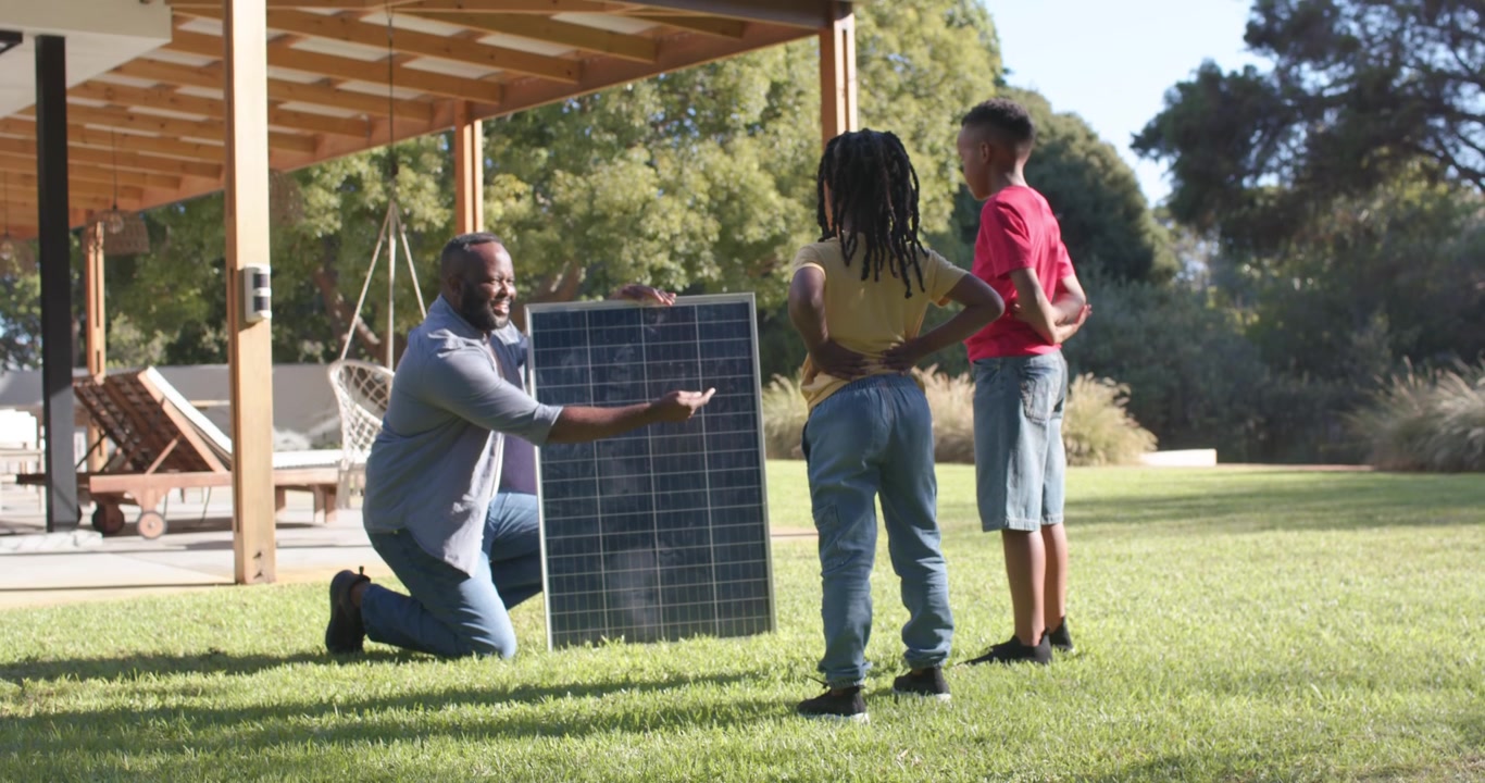 Familiefar knæler, placerer solpanel og peger, underviser to unge mænd på baghavens græsplæne.