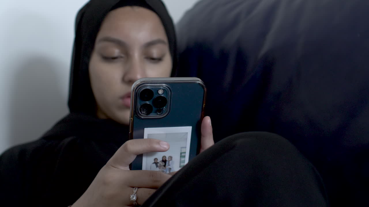 소파에 앉아 있는 동안 히잡을 쓰고 스마트폰으로 타이핑하는 젊은 이슬람 여성들의 배경이 흐릿하다