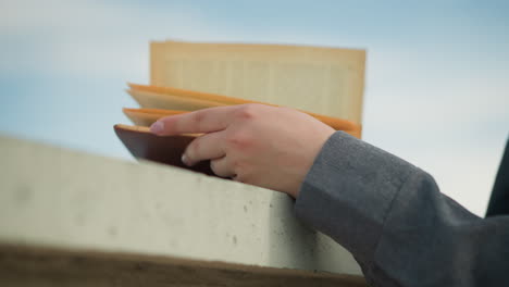 close up hand view of an individual holding a book on a fence as the wind gently blows through the pages, the hand is resting on the book while the wind flips the pages