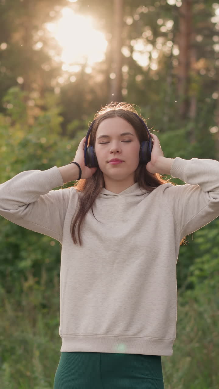 дама с наушниками стоит в лесу. молодая женщина с длинными распущенными волосами наслаждается ритмами релаксационной музыки на свежем воздухе при заходе солнца. вечером расслабьтесь