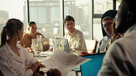 an office worker with black skin color in a white shirt tells his business plan to colleagues at the office table in a room with large windows