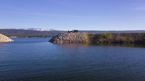 fisherman sits on dyke with parked car behind him, drinking beer, waiting for fish to bite, sunny summer day, aerial shot forward