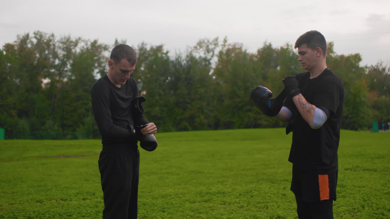 Due uomini bianchi che si scambiano guantoni da boxe, allenamento al crepuscolo su un campo erboso, abbigliamento sportivo casual, dinamica allenatore-allievo, regolazione delle fasce per le mani, incoraggiamento amichevole, preparazione a un leggero sparring, albero panoramico