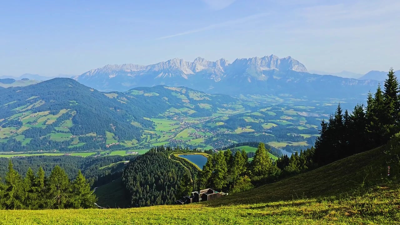 Pemandangan indah dari atas Kitzbuhel, Austria, menampilkan lereng ski hijau cerah, bukit berhutan, kota lembah, danau alpine, dan Pegunungan Alpen Tyrol yang dramatis pada hari perjalanan musim panas yang cerah di Pegunungan Alpen.