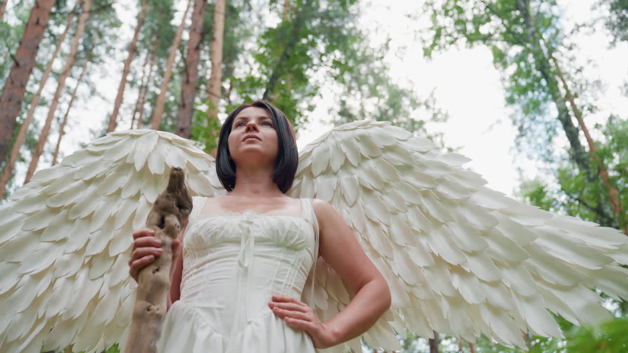 Powerful forest fairy woman with wide white wings and flowing white dress stands proudly among towering trees, exuding mystical energy and strength in magical woodland
