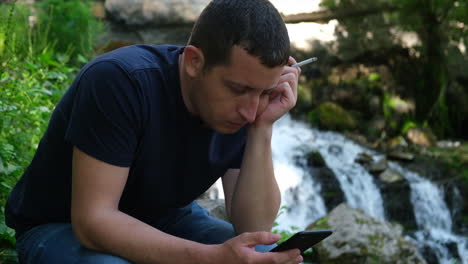 Young-man-holding-a-cigarette-in-one-hand-and-browsing-social-media-on-a-phone-near-a-waterfall