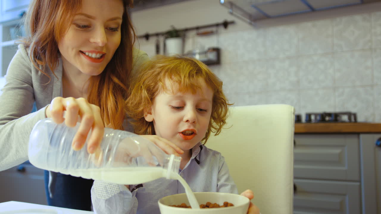 hermosa mujer joven con cabello rojo vertiendo leche en un plato con cereal para su hijo en la cocina