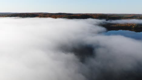 aerial close up of fog above body of water, colourful autumn trees in the background, during cool, bright, sunny day