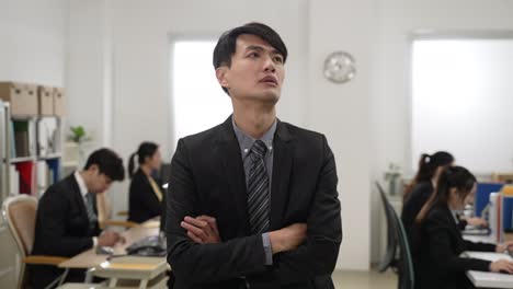 half length portrait of stressed asian team leader worrying about a problem and rubbing head with folded arms as other workers are working at background in the office