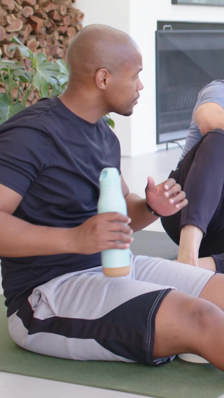 Vertical video: Leaning man speaking at home, black-tee man on green mat grabbing bottle to drink