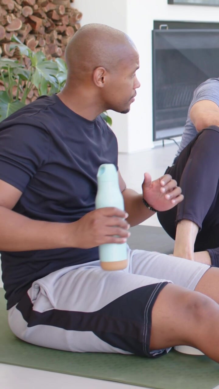 Vertical video: Leaning man speaking at home, black-tee man on green mat grabbing bottle to drink