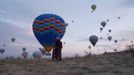 Chica-Vestida-Caminando-En-Un-Campo-Con-Vistas-A-Hermosos-Globos-Aerostáticos-En-Vuelo-En-Capadocia---Toma-Panorámica