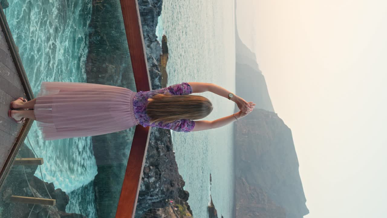 Vertical shot of young woman on vacation raises arms admire sea from terrace