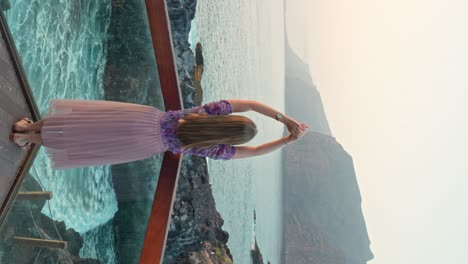 vertical shot of young woman on vacation raises arms admire sea from terrace