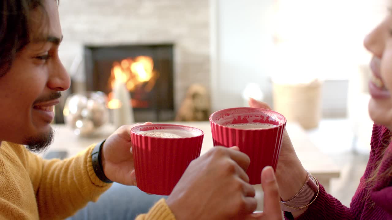 Después de acomodarse en casa, una pareja choca tazas rojas y sorbe latte frente a la chimenea para un momento acogedor