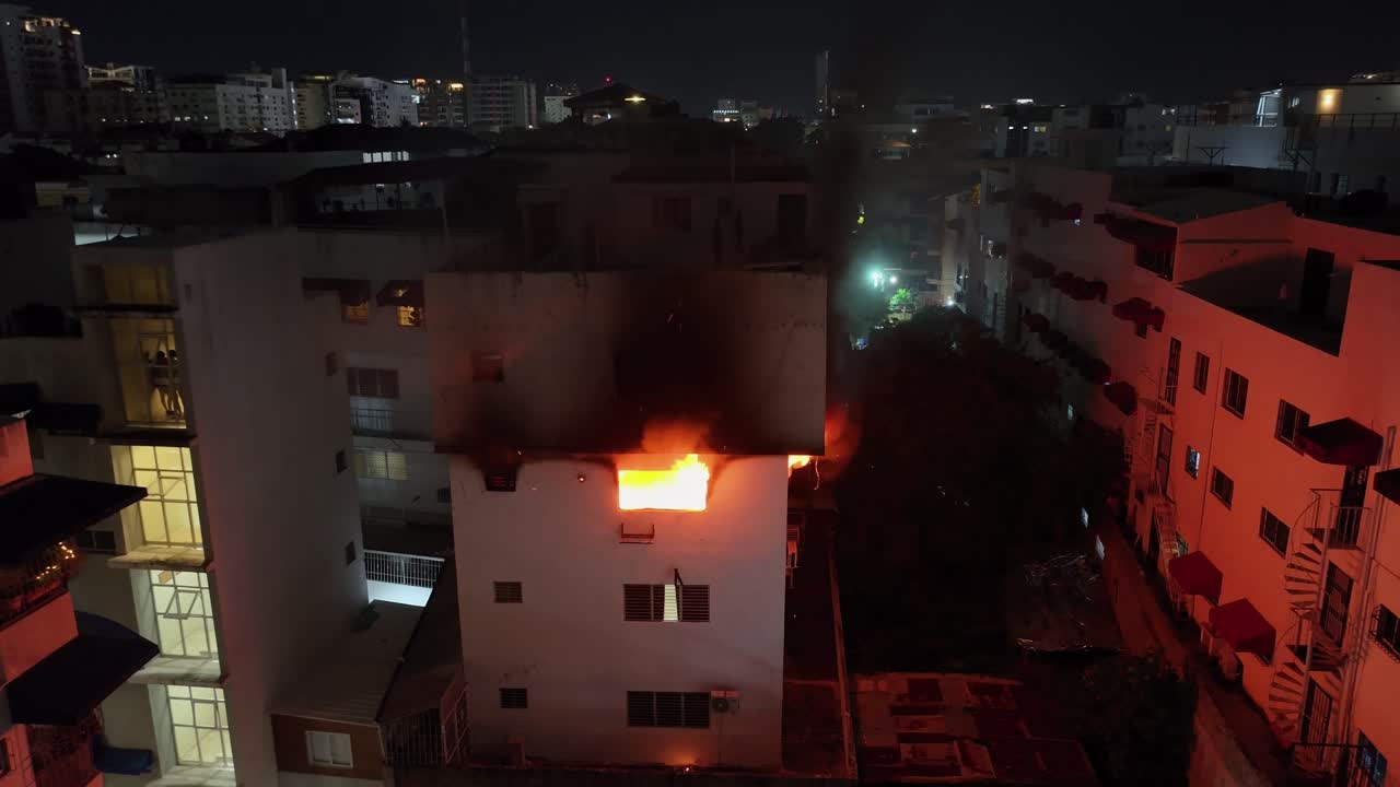 Dangerous burning flames inside apartment of housing area at night. Toxic black smoke rising out of window. Aerial view.