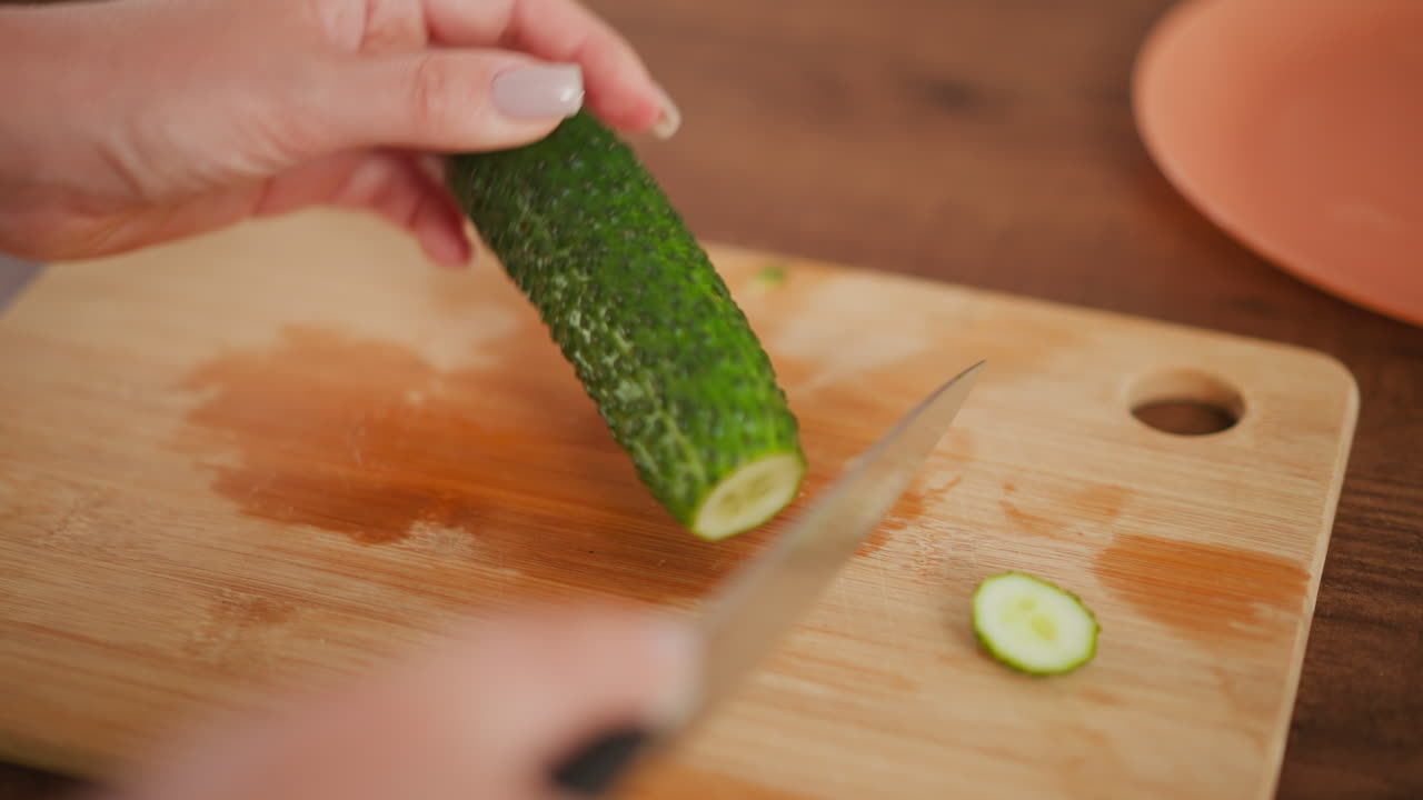 Close up of person holding cucumber firmly on wooden board while slicing off tip with sharp knife, preparing to cut vegetable vertically, with soft lighting and minimal kitchen setup in background