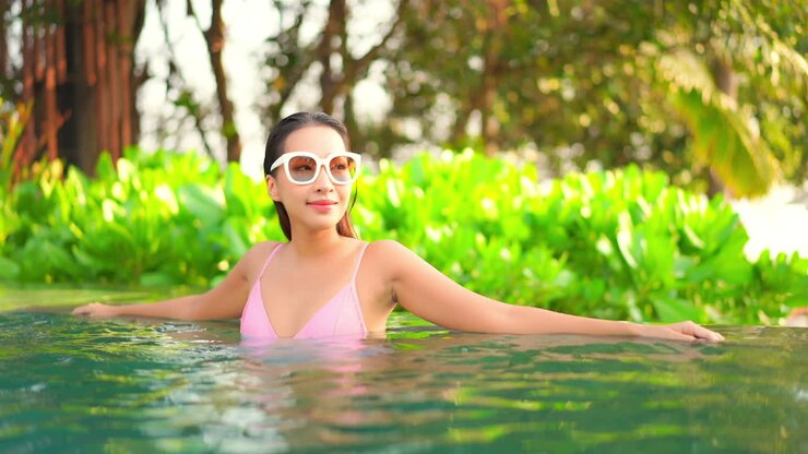 Pretty Asian woman wearing white sunglasses and bathing suit relaxing inside a pool leaning over the edge