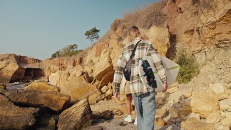 vista trasera de un tipo rubio en una camisa a cuadros sosteniendo una tabla de surf blanca en sus manos y caminando con su novia en una sudadera blanca que también está sosteniendo un tablero de surf de madera amarillo en una playa de arena rocosa por la mañana