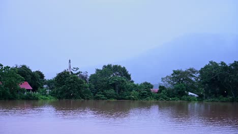 Buddhist-Temple-and-local-farm-community-in-Thailand-flooded-as-a-small-flock-of-Egrets-fly-over-the-flood-water,-a-mountain-at-the-background-covered-by-a-thick-fog-and-clouds