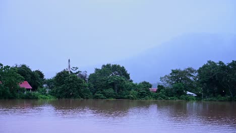 buddhist temple and local farm community in thailand flooded as a small flock of egrets fly over the flood water, a mountain at the background covered by a thick fog and clouds