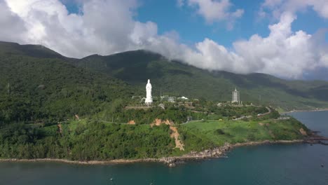 Aerial-view-of-tall-Lady-Buddha-statue-and-temples-with-huge-mountains-and-ocean-in-Da-Nang,-Vietnam