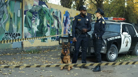 Gemischte-Rassen-Polizist-Und-Polizistin-In-Uniformen-Stehen-Und-Sprechen-Am-Tatort-Mit-Einem-Hund-Am-Polizeiauto