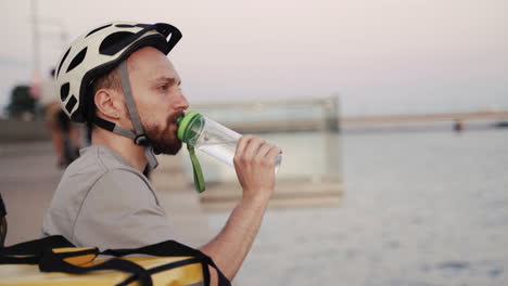 der essenslieferant, der bei sonnenuntergang neben einem fluss sitzt, trinkt während seiner pause etwas wasser