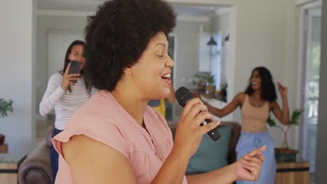 Happy-diverse-female-friends-singing-into-microphone-in-living-room