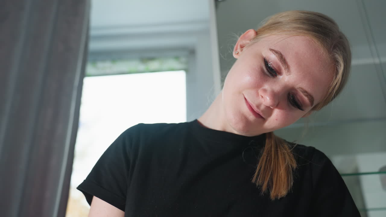 Close up of massage therapist with blonde ponytail wearing black shirt, smiling softly and concentrating on her work, captured with blurred background showing curtain and cabinet in naturally lit room