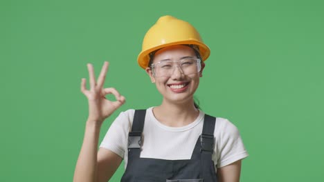 close up of asian woman worker wearing goggles and safety helmet smiling and showing okay gesture to camera while standing in the green screen background studio