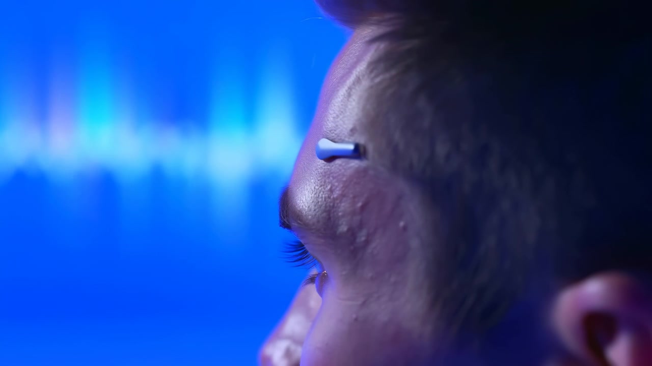 Primo piano ravvicinato del viso mostra un occhio calmo e una tempia con un attacco metallico compatto sulla pelle, mentre morbide fasce di luce ciano e blu rimangono sfocate sullo sfondo.