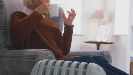 senior woman with hot drink keeping warm by portable radiator at home in cost of living crisis