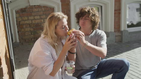 young couple sharing a delicious hamburger and drinking a cold drink together, sitting on the sidewalk